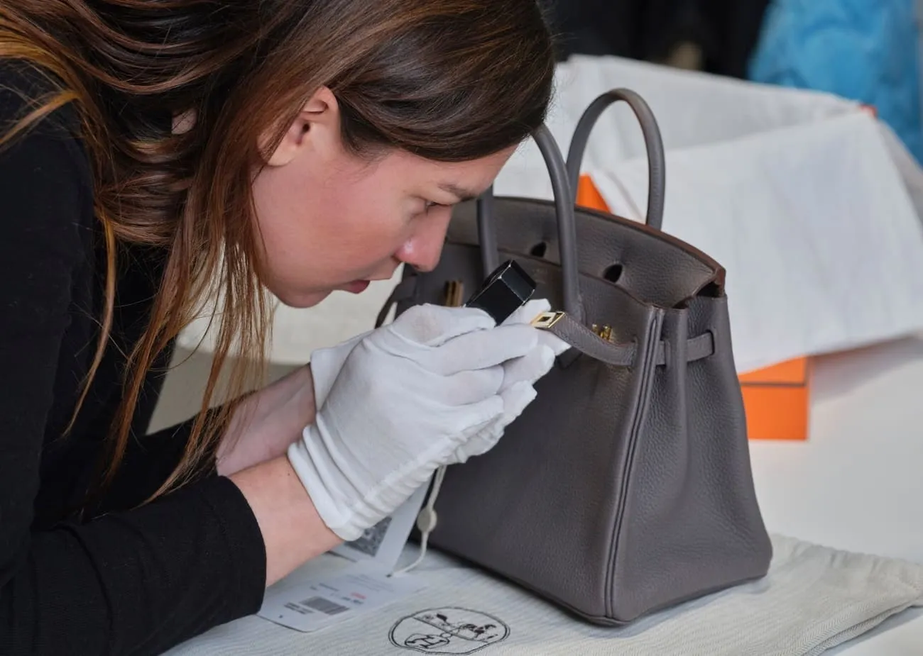 Specialist in white gloves inspecting the hardware of a leather handbag with a loupe
